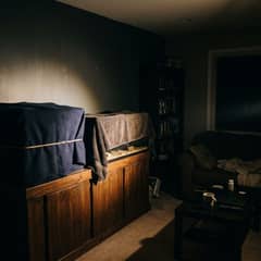 A dark living room during a power outage lit by a single flashlight, an aquarium wrapped in a blanket on its stand and a towel-covered reptile enclosure beside it